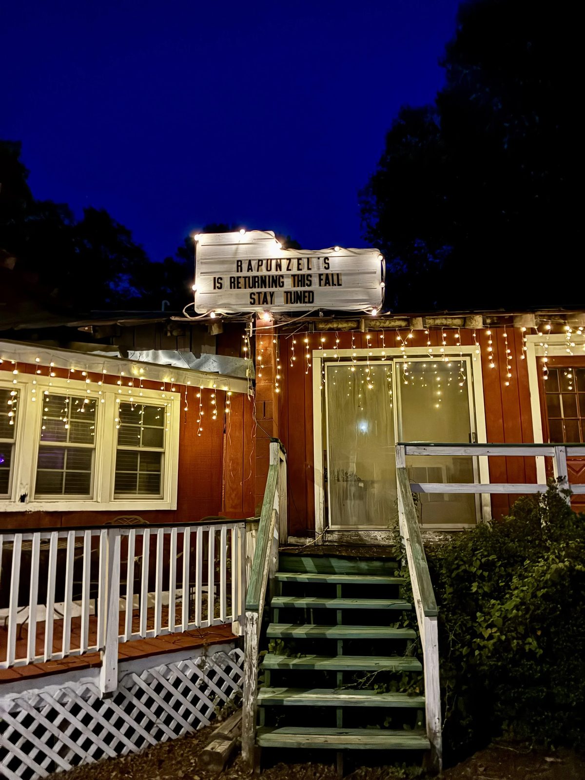 Rapunzel's at dusk: the white letterboard marquee above the barn-red building reads RAPUNZEL'S IS RETURNING THIS FALL — STAY TUNED, under strings of warm lights.